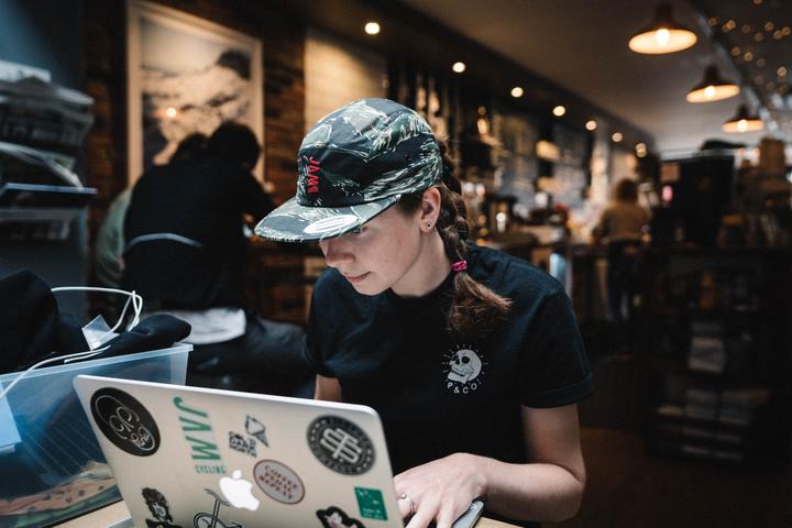 Jamouflage 5 panel cap showcasing HOT PINK am logo symbol worn by JAM female employee Jess sitting working at laptop in a cafe