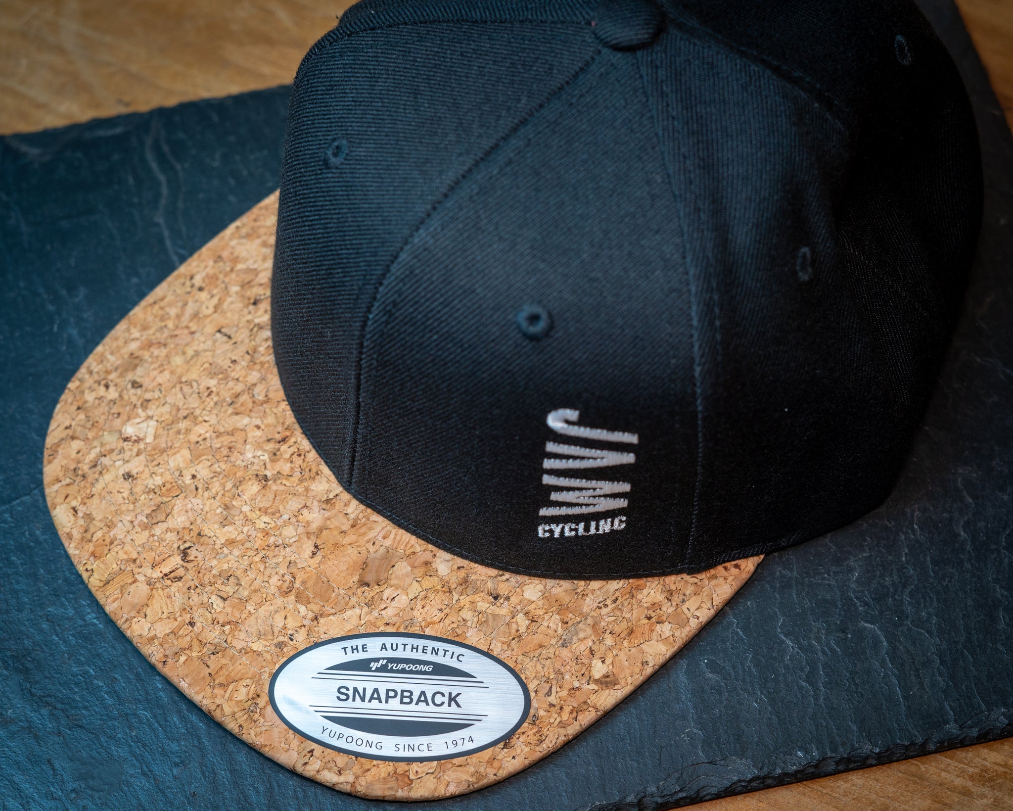 JAM snap back cap featuring cork peak stitched JAM logo up close detail on a black slate background cool product photography