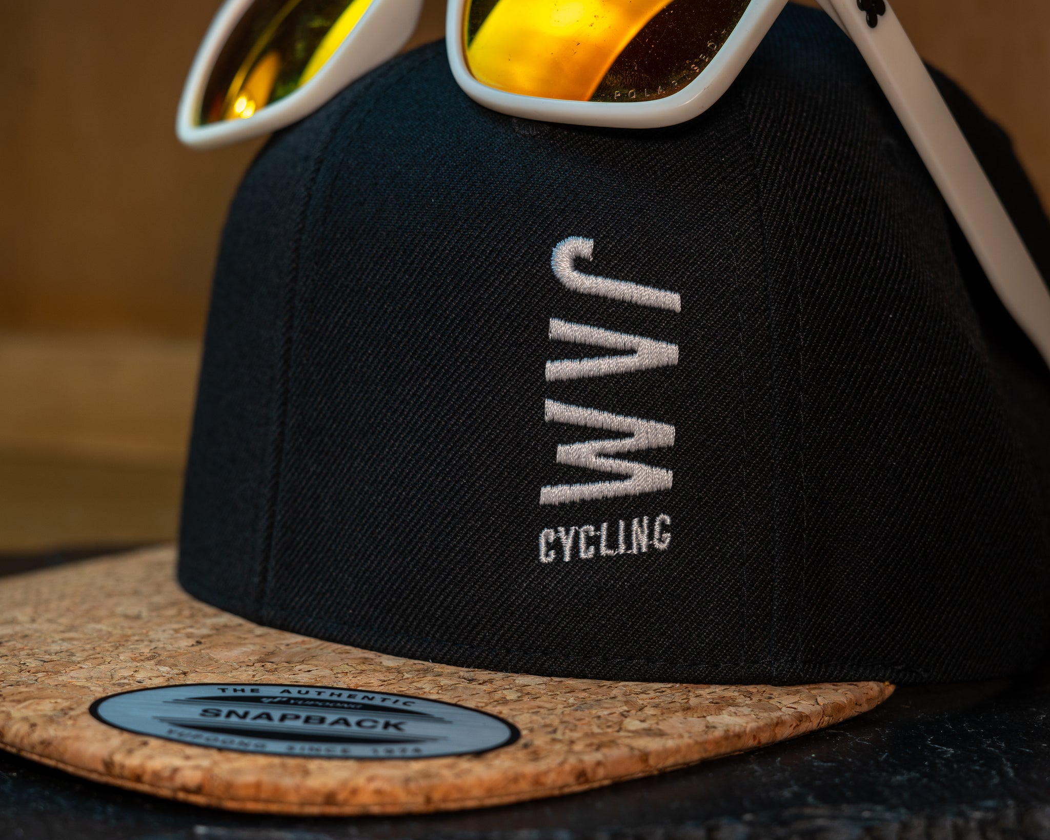 JAM snap back cap featuring sunglasses cork peak stitched JAM logo up close detail on a black slate background cool product photography